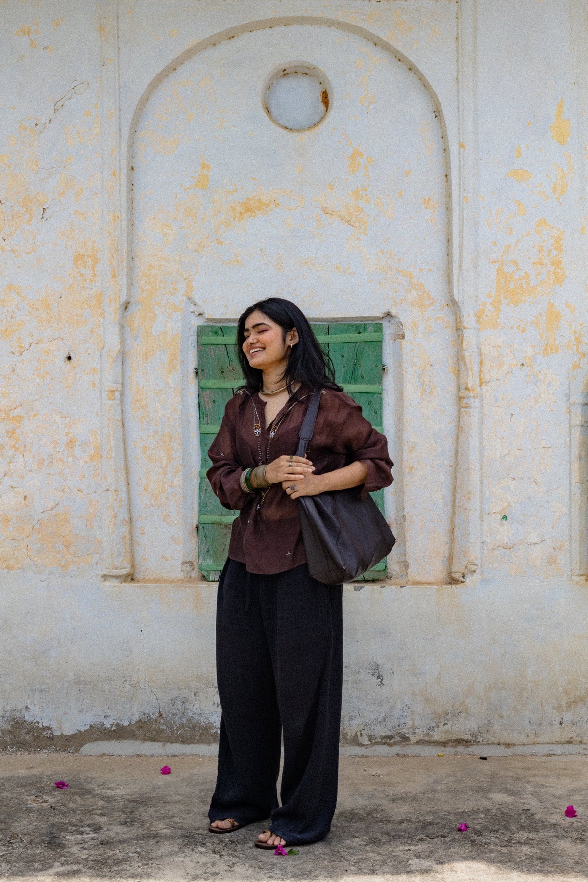 A woman in a side profile wearing a white long-sleeve top, embellished brass sequence, and matching straight pants, standing in a minimalist white-walled architectural setting.