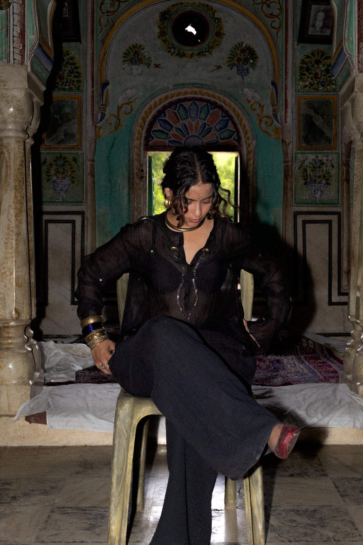 A woman wearing a sheer black long-sleeve kurta and black pants sits on a stool inside a highly ornate, arched room with painted walls and columns.