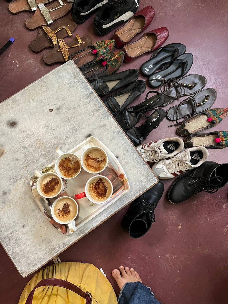 An overhead view of a rustic table holding five cups of coffee, surrounded by numerous shoes and sandals on a reddish-brown floor. edge.
