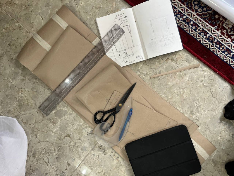 Some patterns laid out on a marble floor, surrounded by sewing and crafting tools including a notebook with sketches, a ruler, scissors, a pen, and a black tablet.