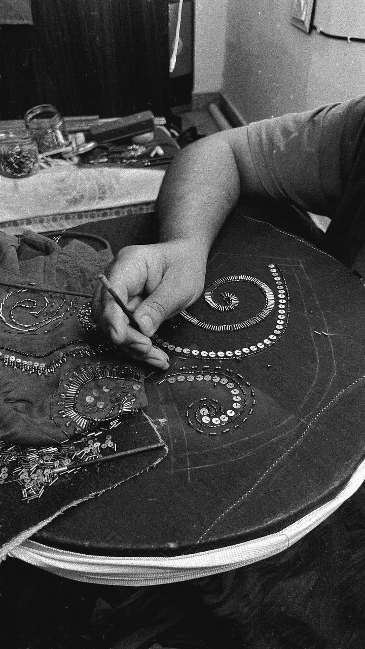 A black and white close-up of a person working on adda, applying metallic embellishments in spiral patterns.