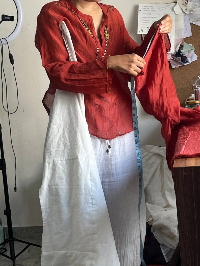 A person in a home studio setting wearing a red embroidered kurta and a white linen pant, measuring them with a blue tape measure. A ring light stands to the left, and a desk with papers is on the right.