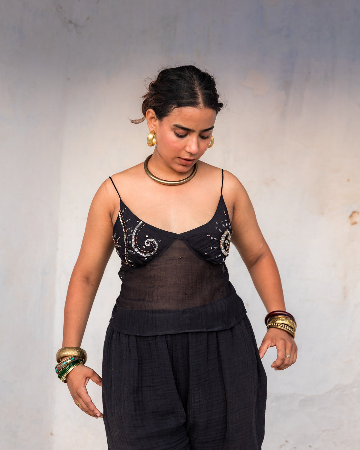 A woman wearing a black sheer embellished camisole top and black wide-leg pants, accessorized with gold jewelry, standing against a plain light gray wall.