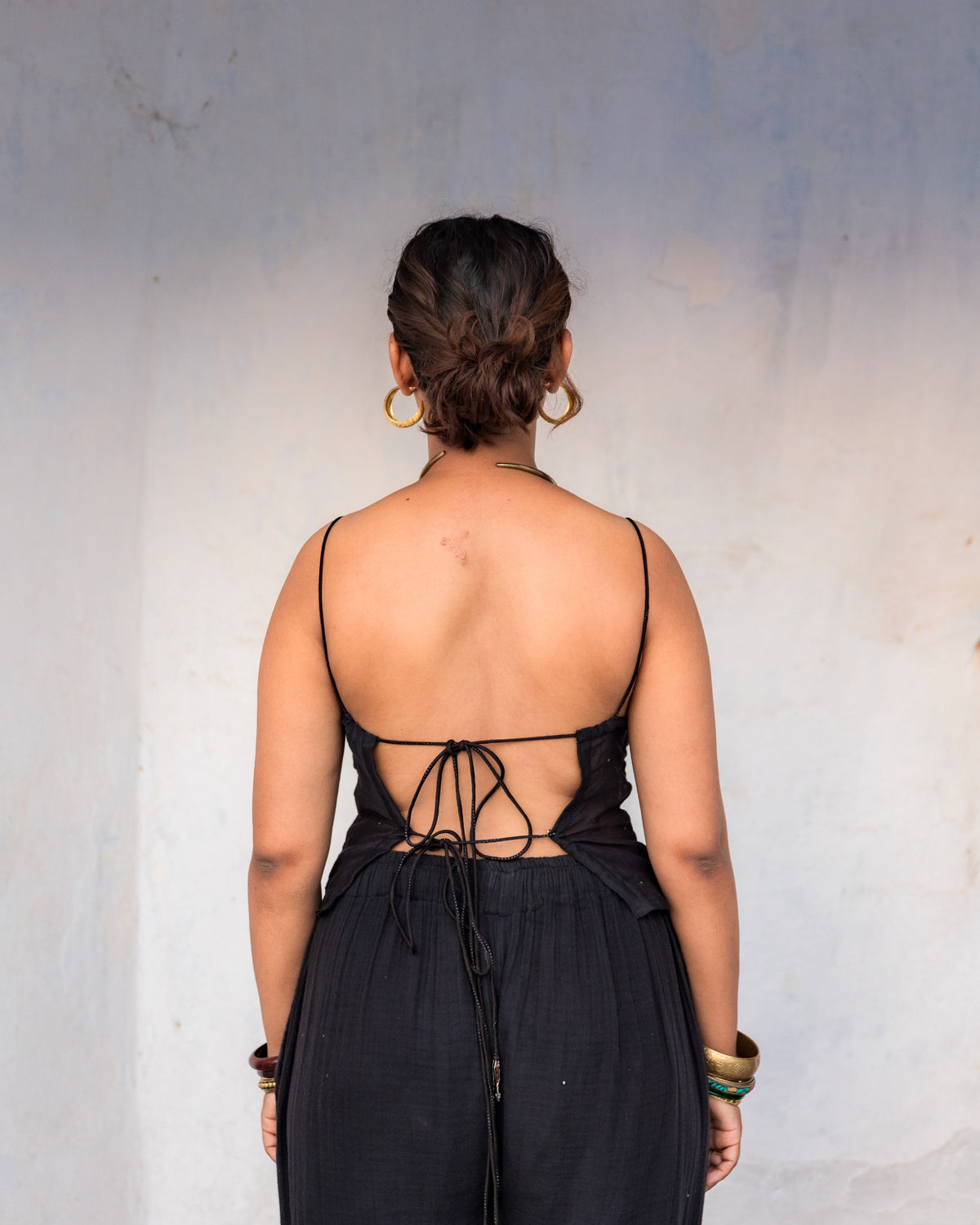 A woman viewed from behind wearing a black open-back top with thin straps and tie details at the back, standing against a plain gray wall.