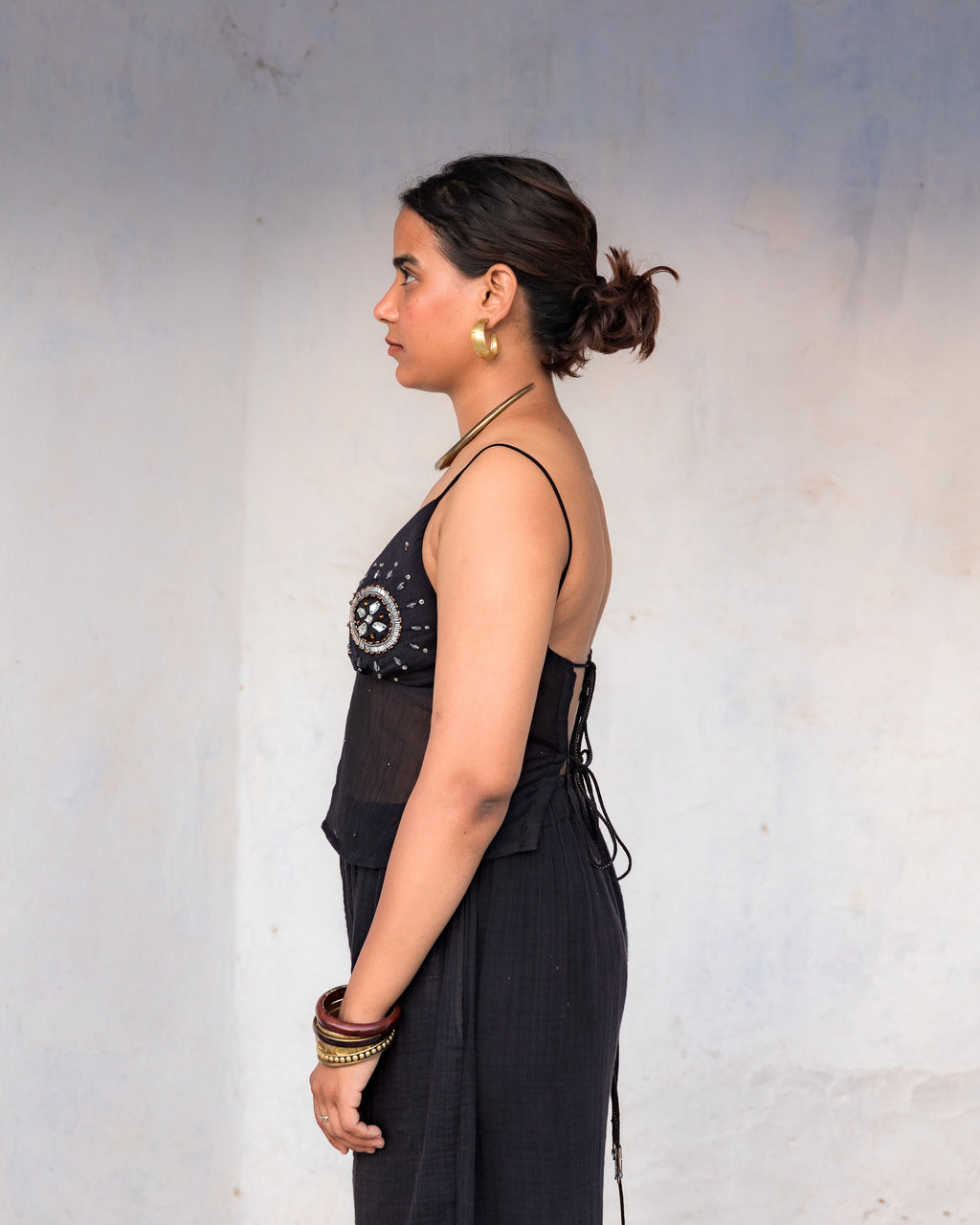A woman in side view wearing a black sleeveless mul chanderi top with a decorative embellished bodice and side tie, standing against a plain light gray wall.