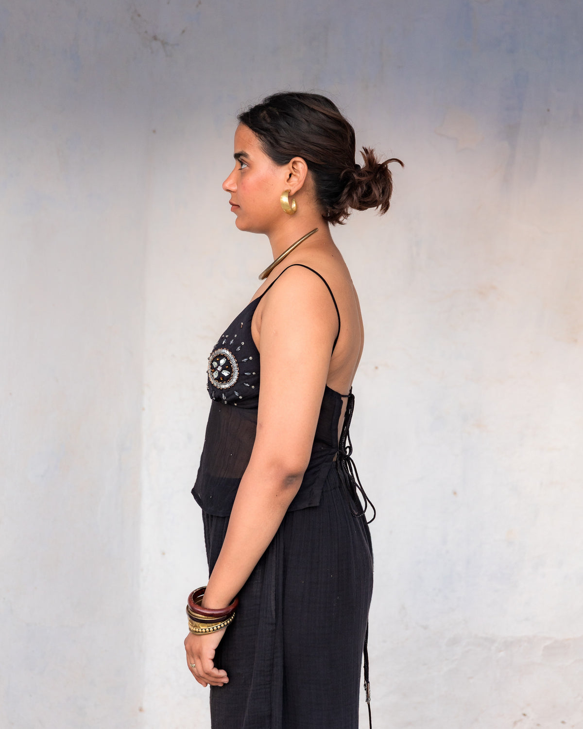 A woman in side view wearing a black sleeveless mul chanderi top with a decorative embellished bodice and side tie, standing against a plain light gray wall.