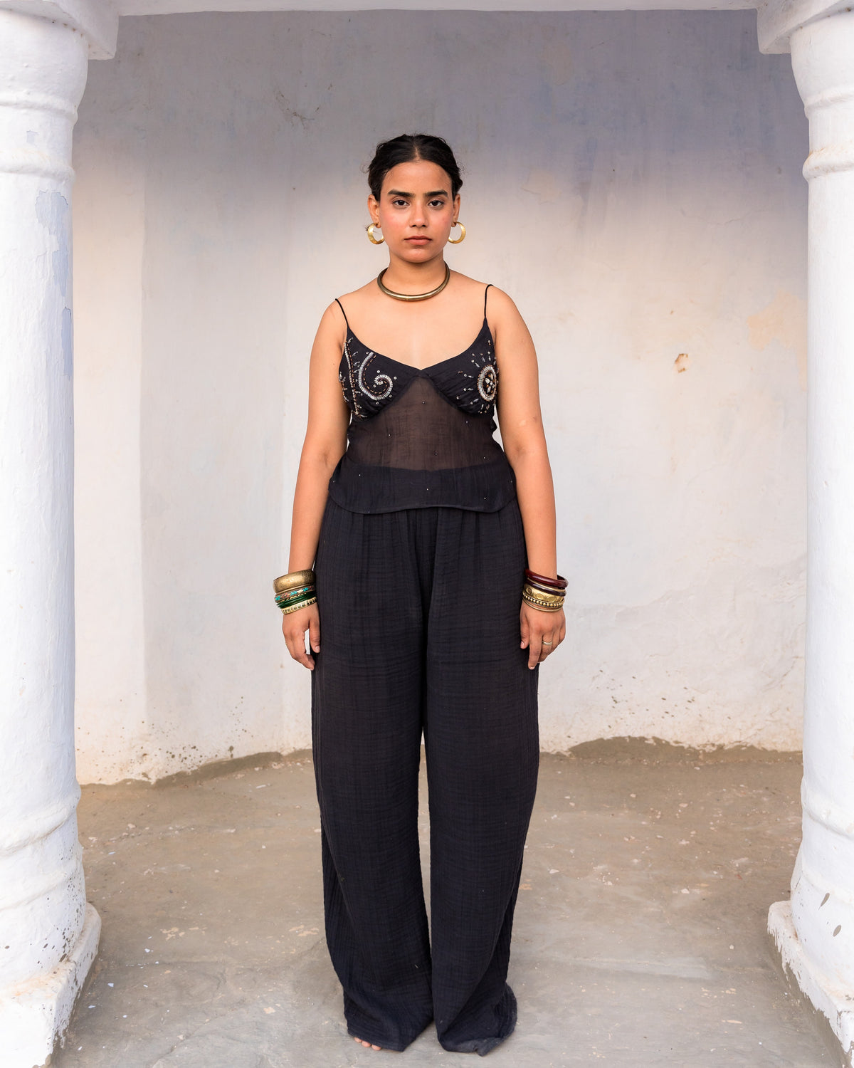 A woman stands in a doorway wearing a black sleeveless dori top with sheer panel and silver embroidered details, paired with loose black pants. She is accessorized with gold jewelry.