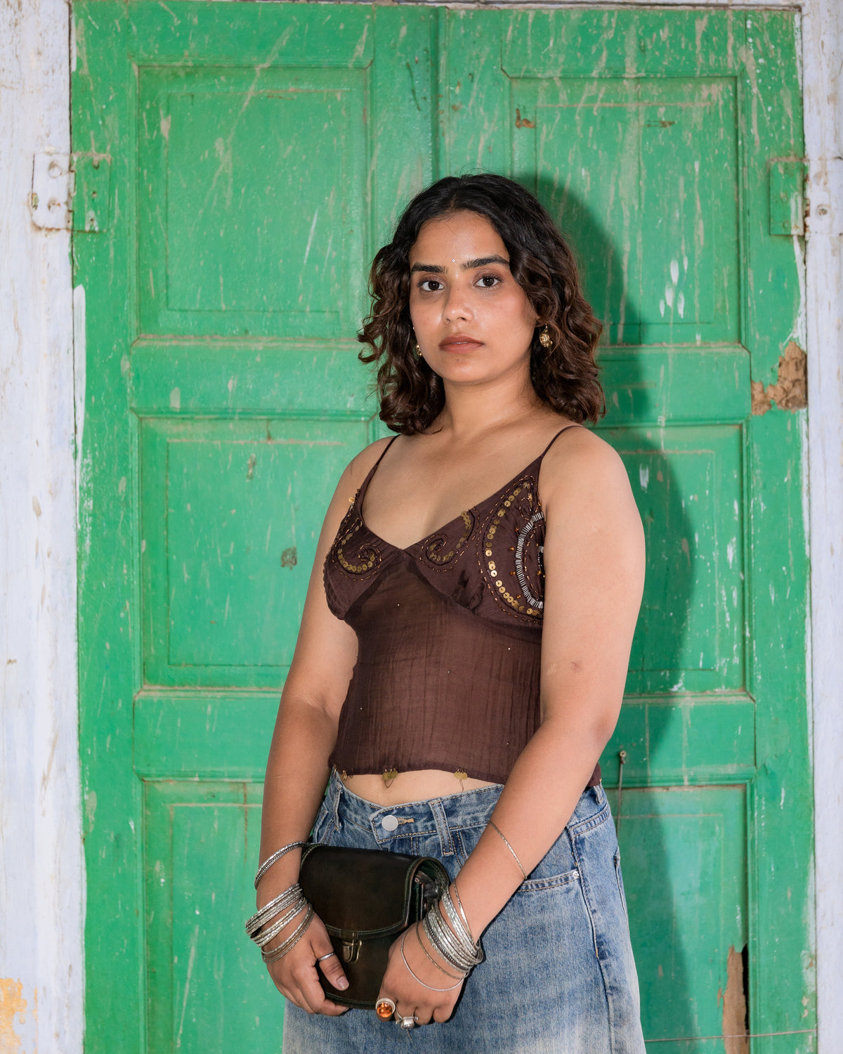 A woman stands in front of a weathered green wooden door, wearing a dark brown embroidered camisole top and a light-wash denim jorts, holding a black clutch bag.