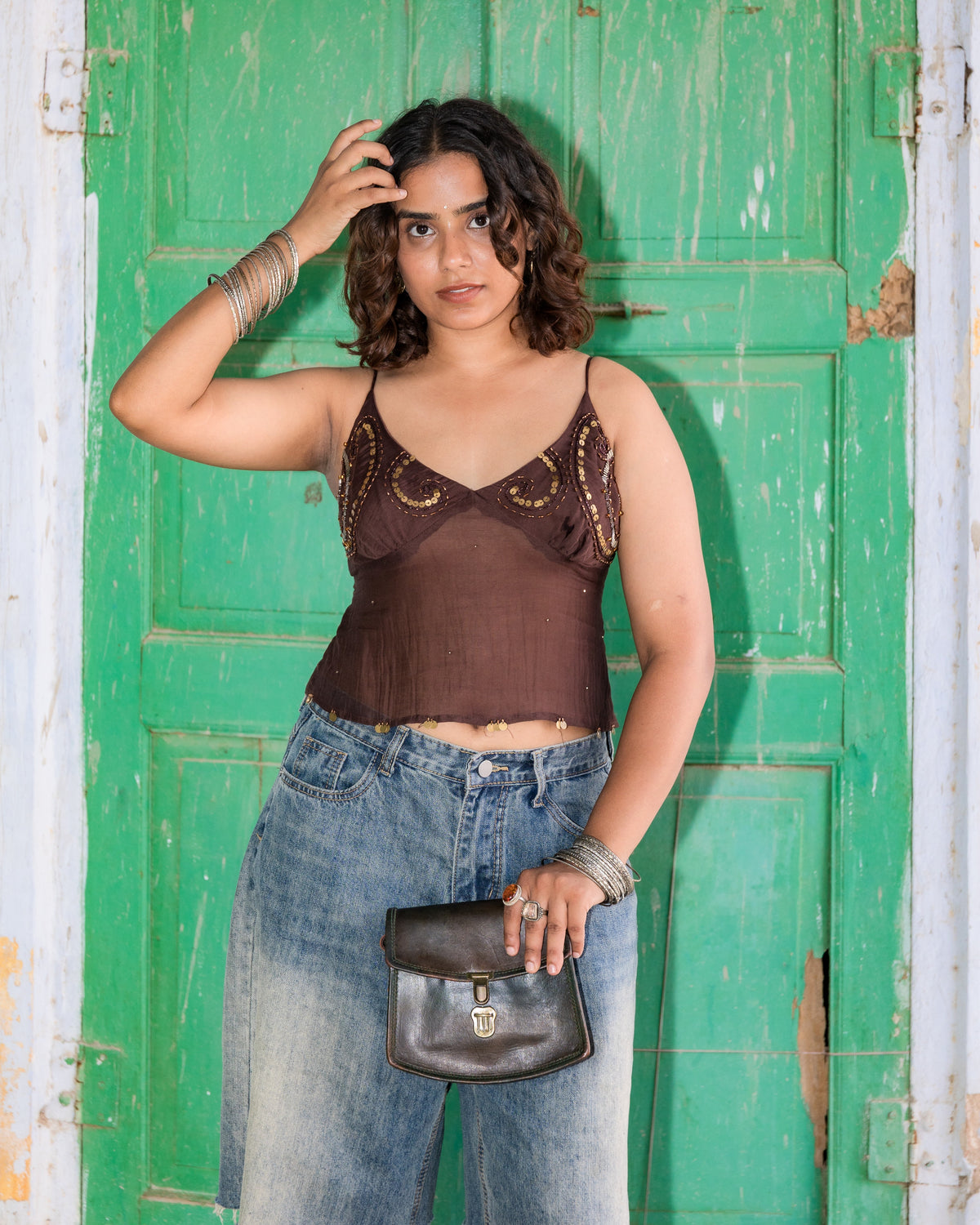 A woman stands in front of a weathered green door wearing a brown spaghetti-strap crop top with  deep neck line gold-tone circular embellishments, blue denim shorts, and holding a small dark brown crossbody bag.