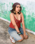 A woman wearing a rust red embellished deep neck string tying top, denim shorts, and white sneakers with red stripes.