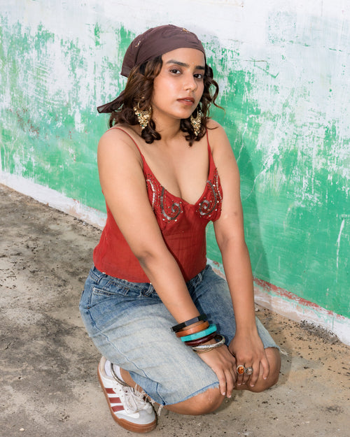 A woman wearing a rust red embellished deep neck string tying top, denim shorts, and white sneakers with red stripes.