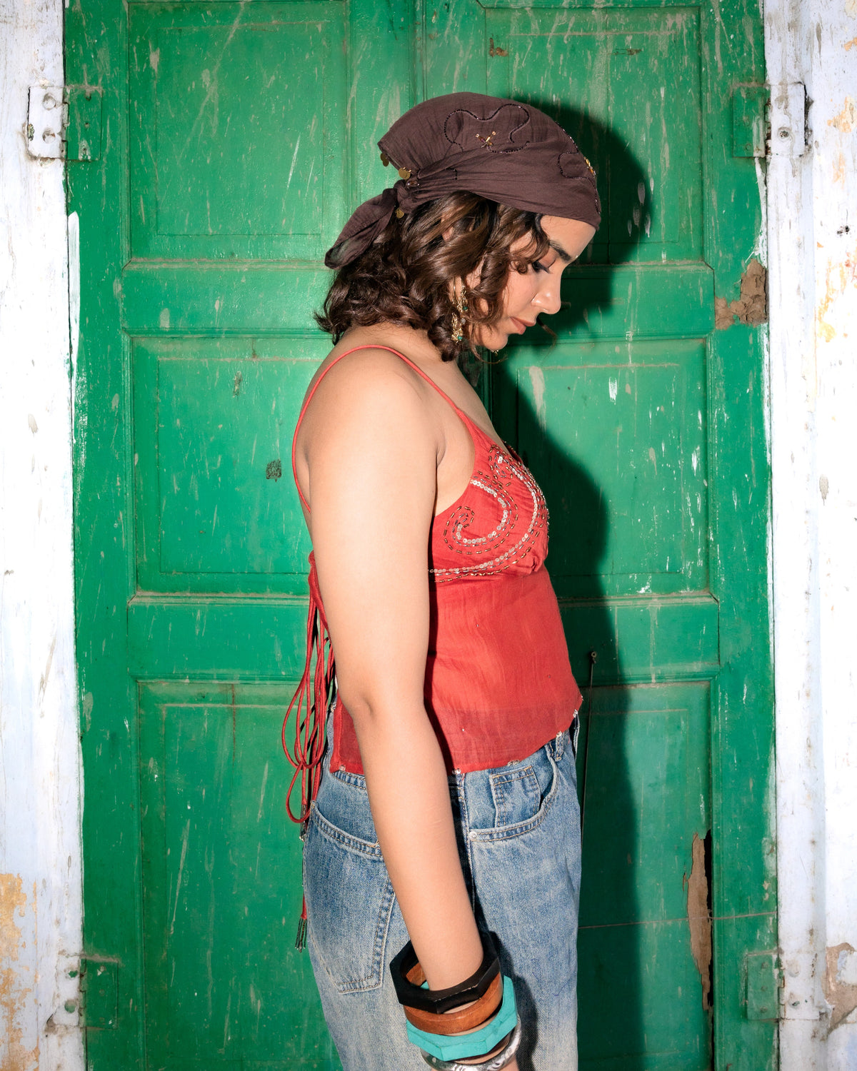 A person in profile wearing a brown bandana, a rust-red cropped top with handdone embroidery, and light-wash blue jorts, standing in front of a green door.