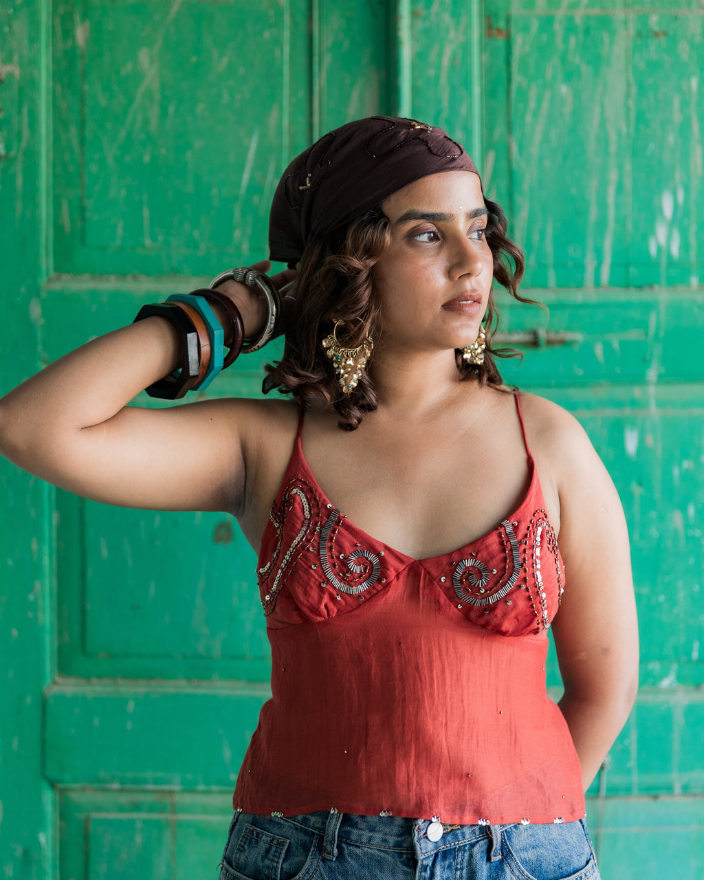 A woman wearing a rust-orange bead embroidered camisole top, blue jeans, and a dark headwrap stands in front of a weathered green wooden door.