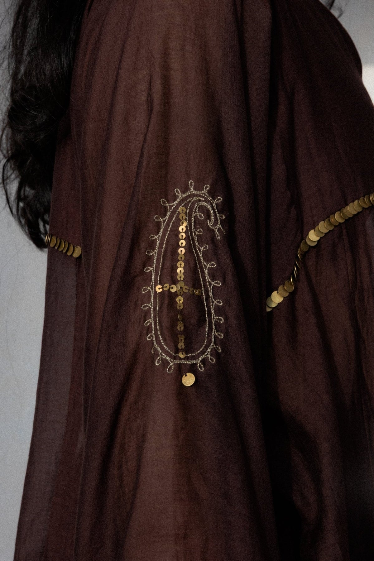 A close-up of a dark brown traditional garment featuring intricate gold sequin embroidery and coin detailing on the sleeve.