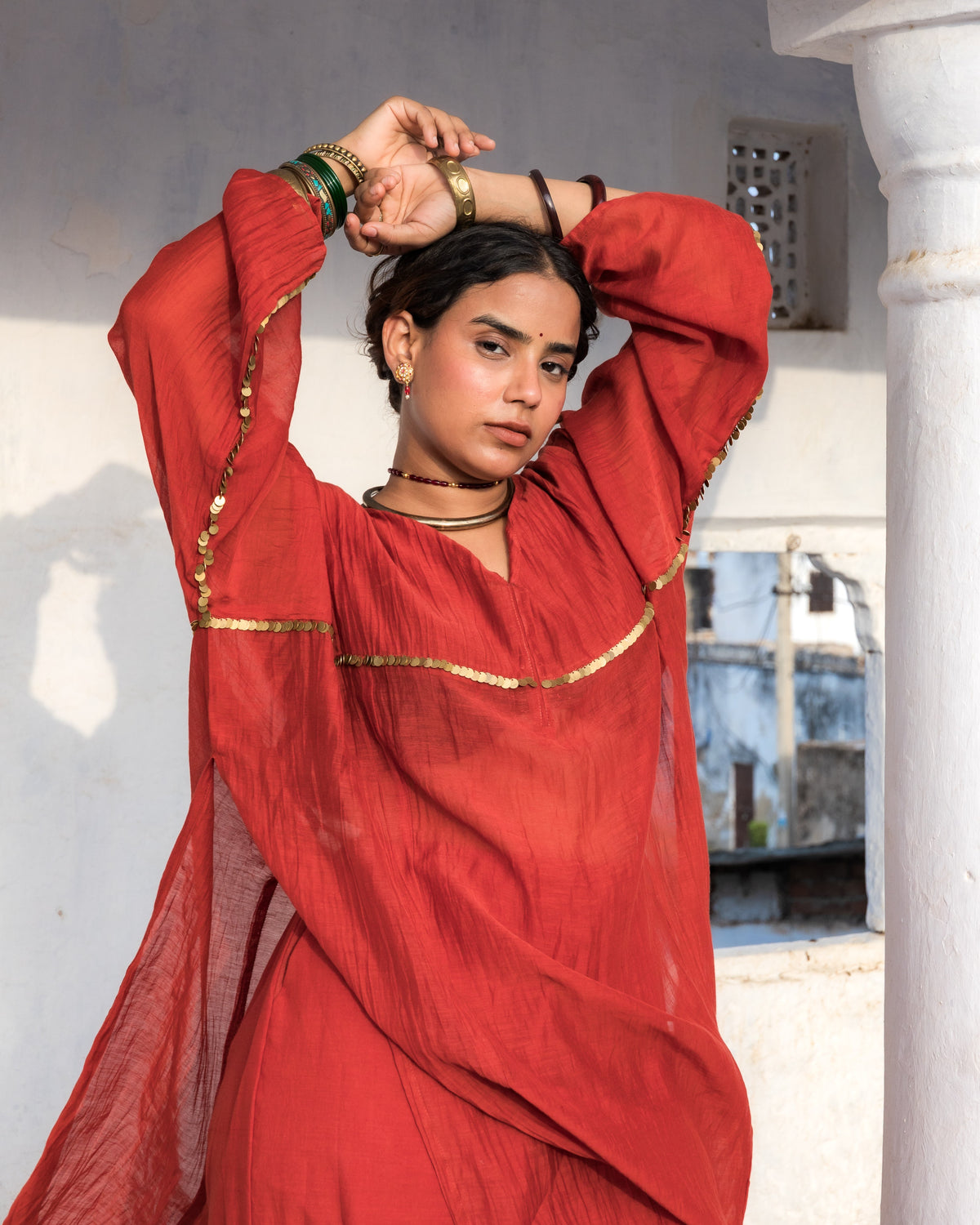 A woman wearing a rust-red traditional outfit with gold metal sequence work  and multiple bangles, posing with her arms raised in front of a white architectural background.