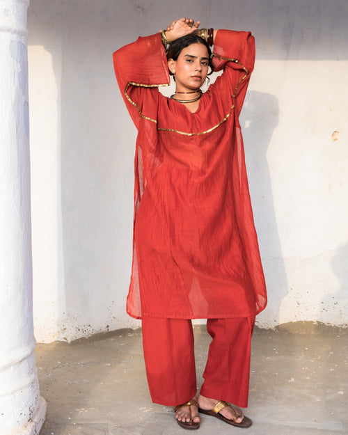 A woman modeling a sheer rust-red traditional kalidar outfit with matching cotton pants with pockets, and brass coin embellished, standing against a plain white wall.