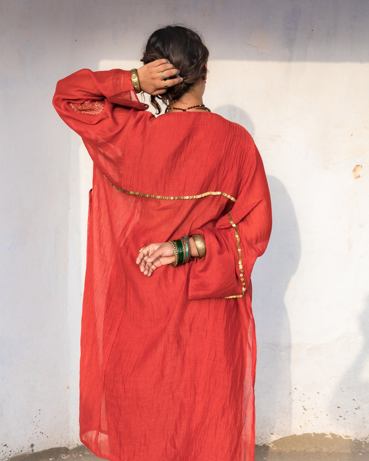 A person viewed from behind wearing a loose, long-sleeved rustic-red kalidar kurta with gold trim details, standing against a plain off-white wall.