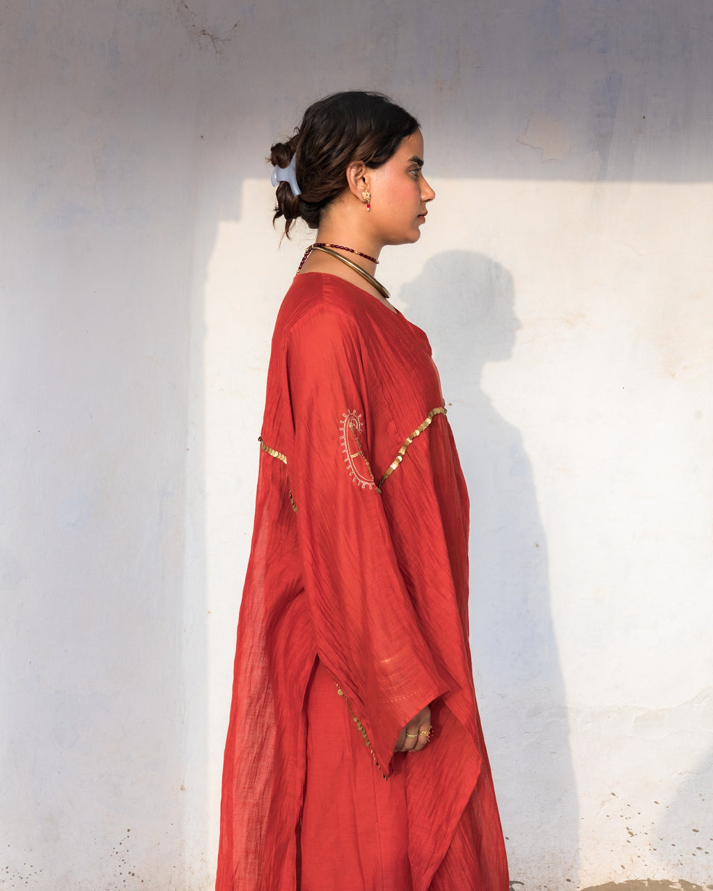 A woman in profile wearing a loose, rust-red kaftan dress set with gold embroidery along the seam line and paisley embroidery on sleeve, standing against a textured off-white wall.