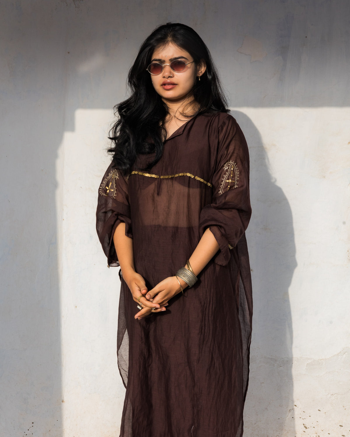 A woman wearing a dark brown sheer tunic with gold embroidery and matching trousers, standing against a textured off-white wall.