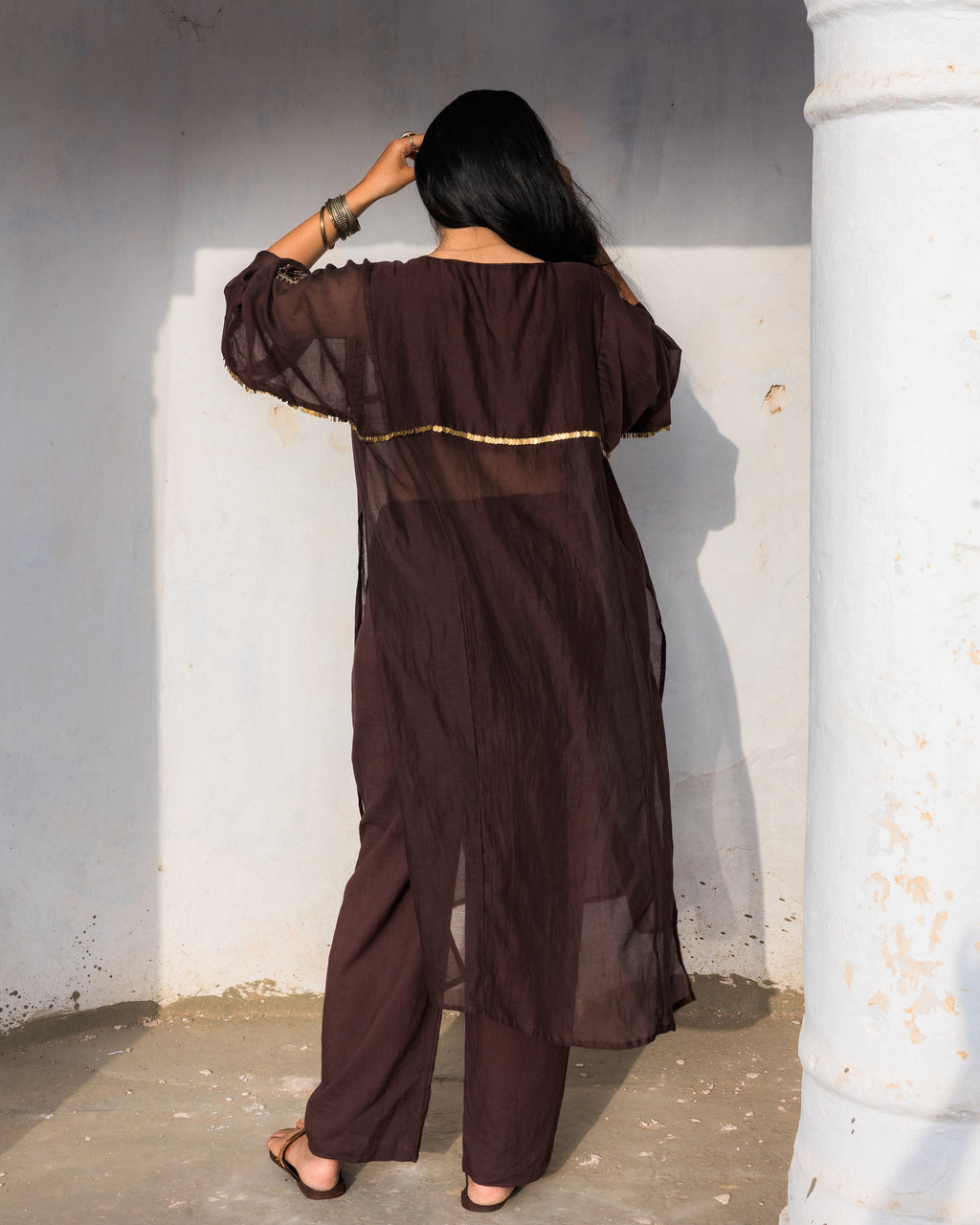 A woman stands with her back to the camera wearing a sheer dark brown long kurta and narrow cotton  pants with pocket set with a thin gold horizontal stripe of brass sequence across the back and the front. 
