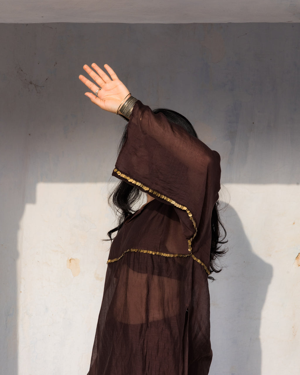 A person wearing a flowing dark oversized brown sheer kurta set with gold brass large sequence, posed with one arm raised against a plain light wall.