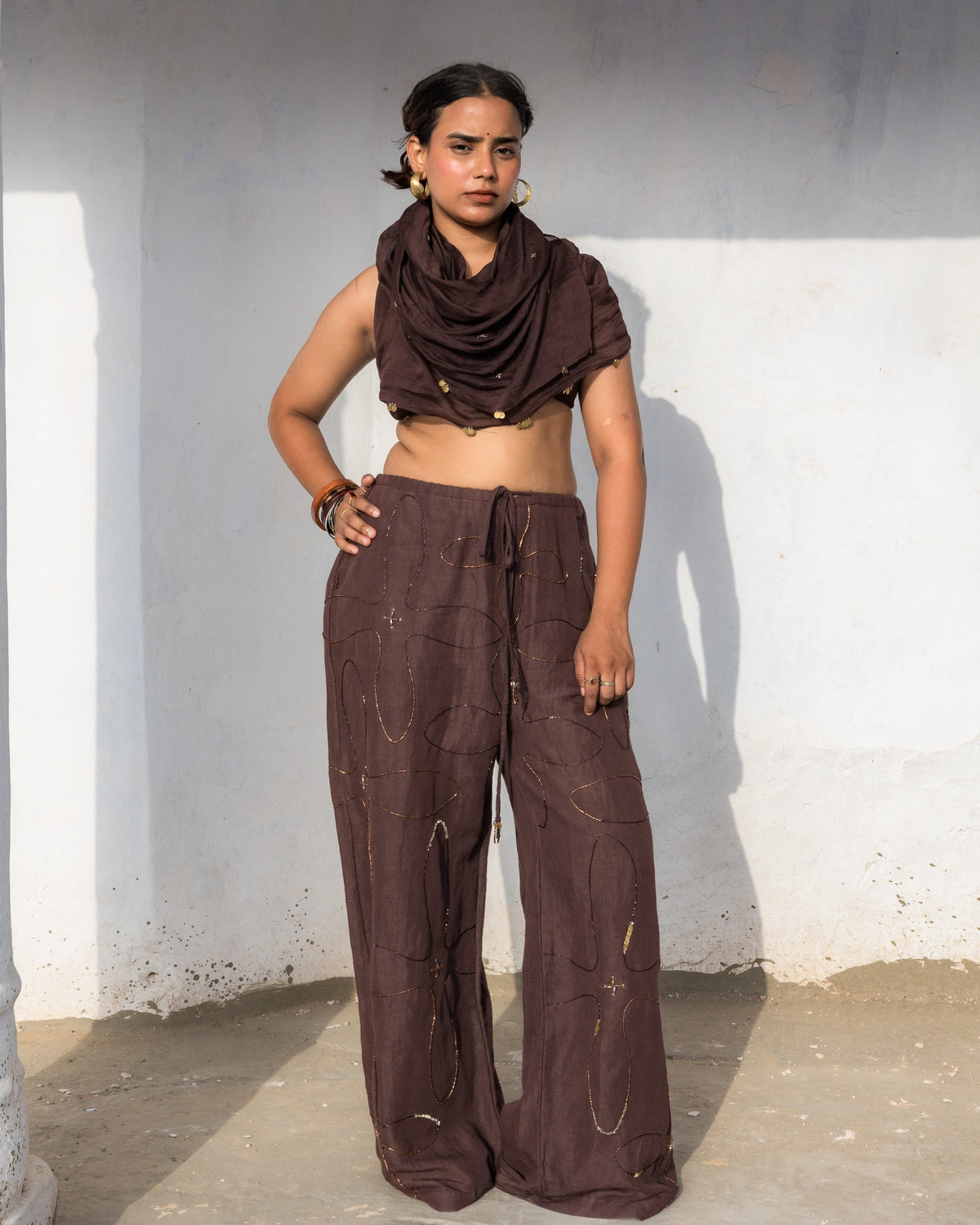A model stands against a white wall wearing a dark brown straight leg loose fitted pants with a drawstring  waist, intricately embellished with sequins and handwork.