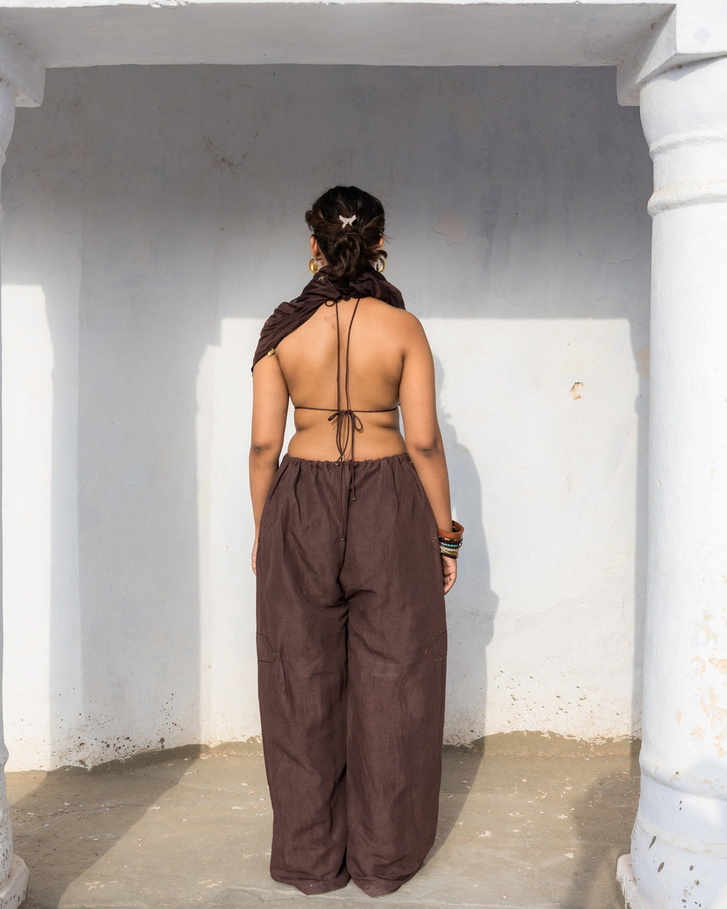 A woman stands with her back to the camera wearing a brown backless wrap top and wide-leg pants in brown linen cotton with handwork in a white-walled corridor.