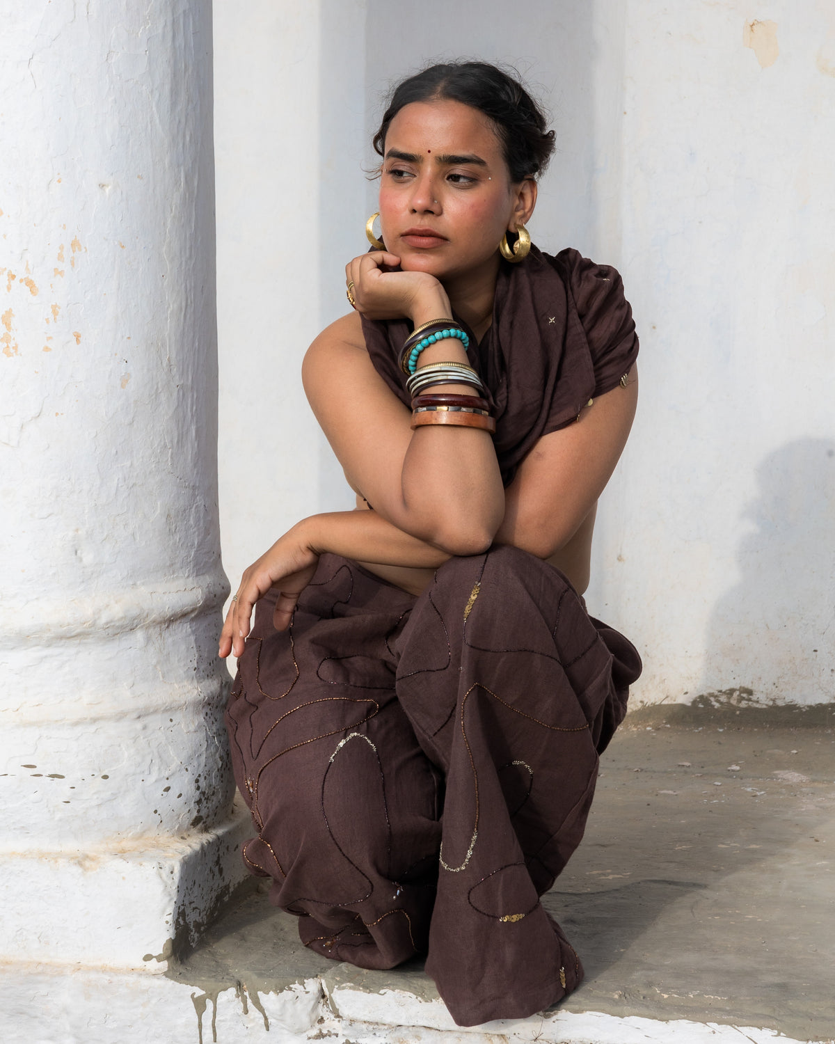 A woman wearing a dark brown linen cotton pants which is uniquely embellished sits on the steps of a white-walled building. She is wearing large gold hoop earrings and multiple bangles on her wrist.
