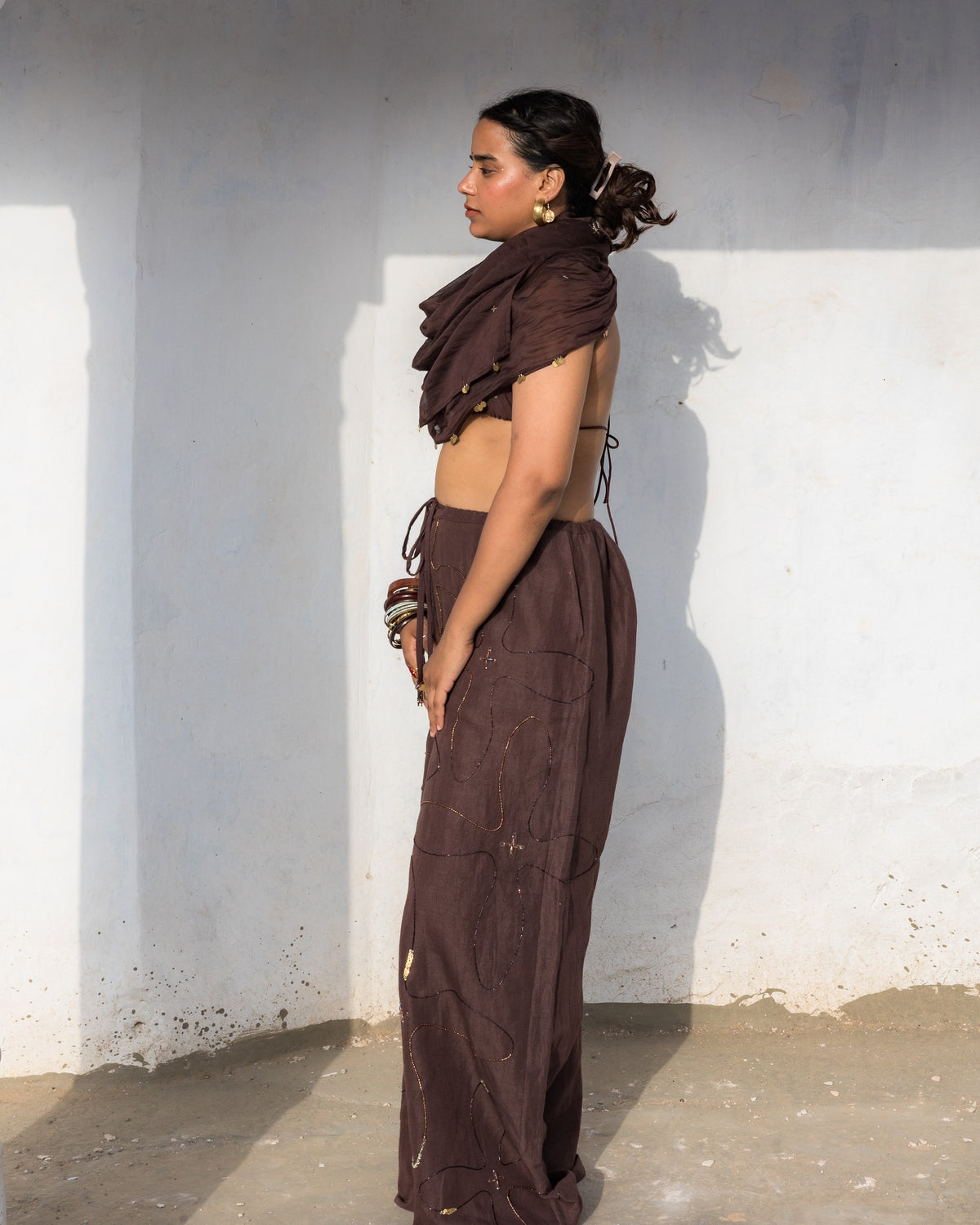 A woman stands in profile against a white wall wearing a dark brown loose pants made of cotton linen and with detailed beadwork.