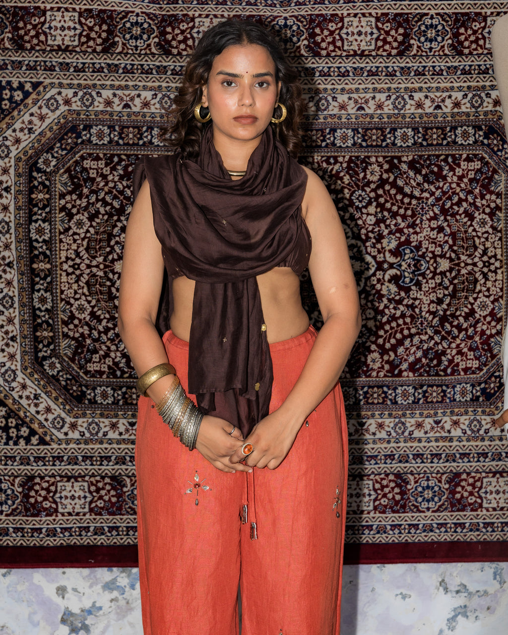 A woman stands in front of a large patterned rug, wearing a brown chanderi dupatta and rust-orange linen pants with modern handbeadwork and silver bangles.