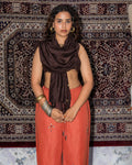 A woman stands in front of a large patterned rug, wearing a brown chanderi dupatta and rust-orange linen pants with modern handbeadwork and silver bangles.