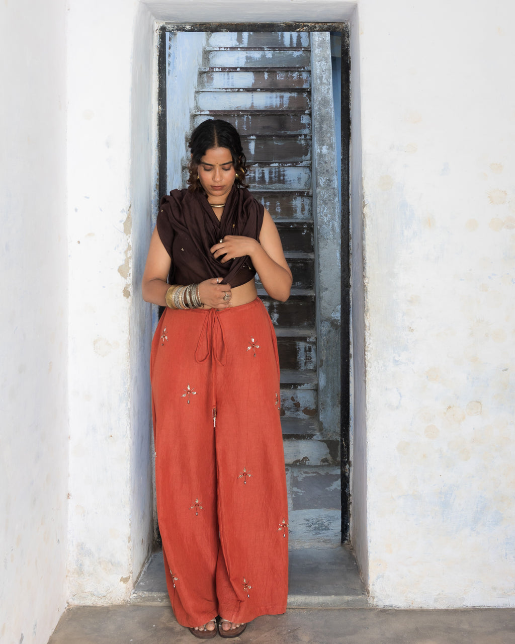 A woman stands in a doorway wearing rust linen pants straight loose fit with embroidered stone details, set against a rustic white wall and concrete floor.