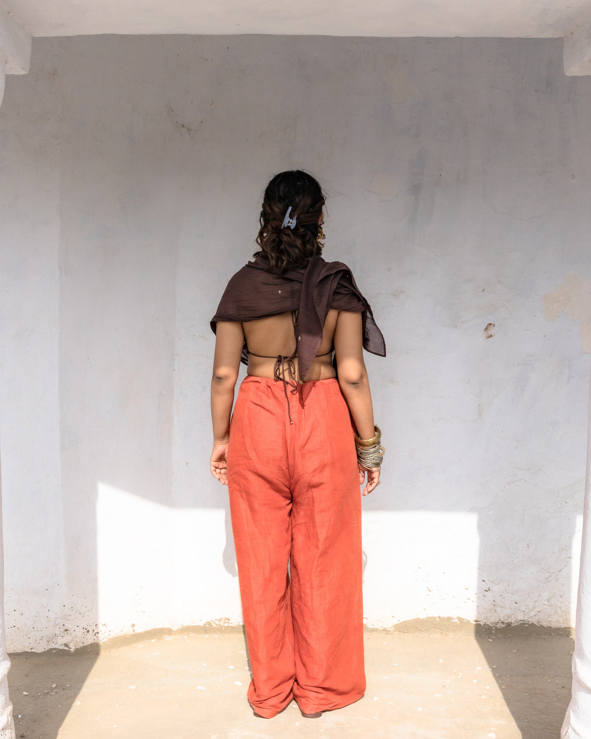 A woman with their back to the camera, wearing rust-colored wide-leg floor touching pants in cotton linen with handwork in front and a chanderi scarf.