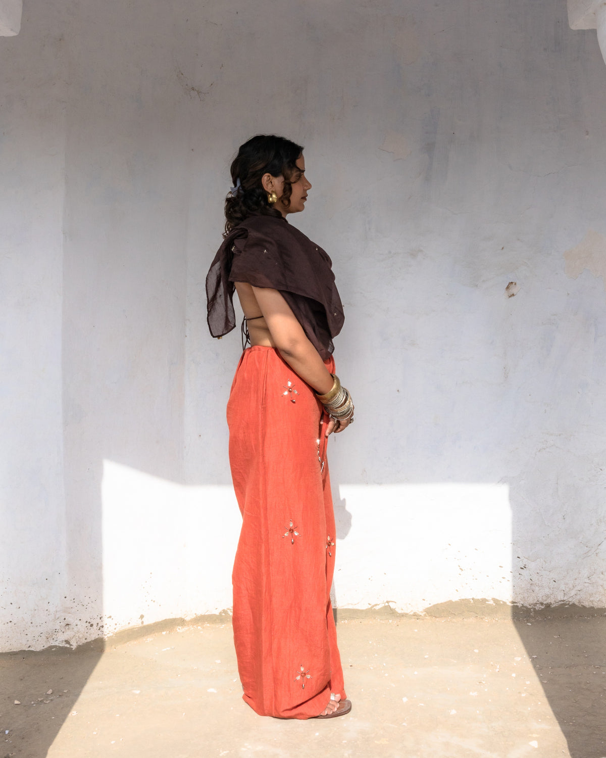 A woman stands in profile inside a white-walled architectural alcove, wearing a long rust-orange linen pant with handwork, small metallic embellishments.