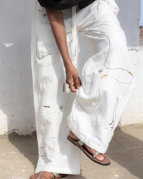 A person wearing loose white drawstring pants with abstract gold and silver line embroidery and a black sheer scarf, standing barefoot in silver brown sandals against a white wall.
