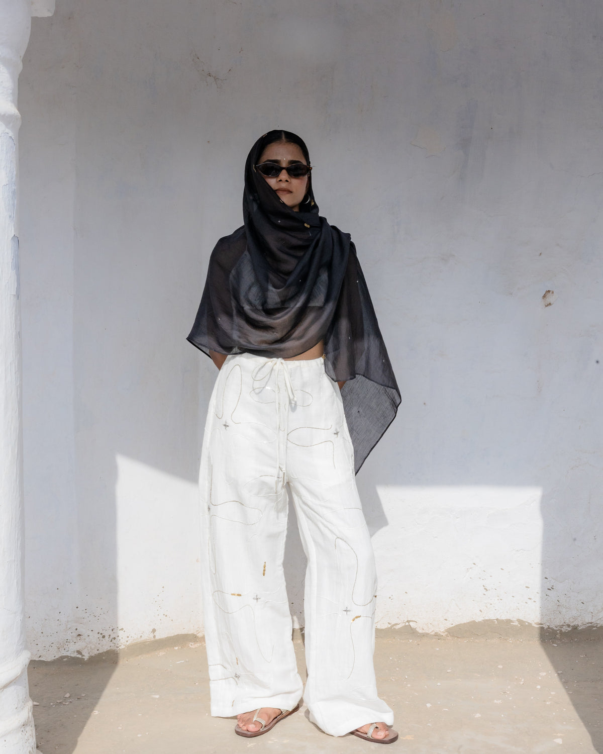 A person standing in a white-walled architectural setting, wearing a black draped headscarf and loose white straight-leg pants with detailed gold and silver beadwork, and sunglasses.