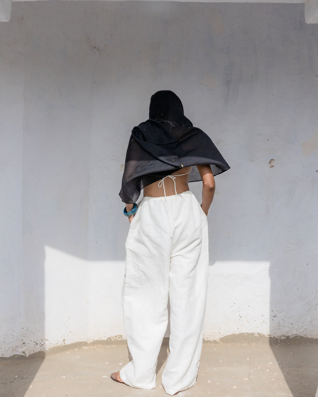 A person standing with their back to the camera in a minimalist white-walled room, wearing a loose white linen cotton trousers with drawstring  and handwork.