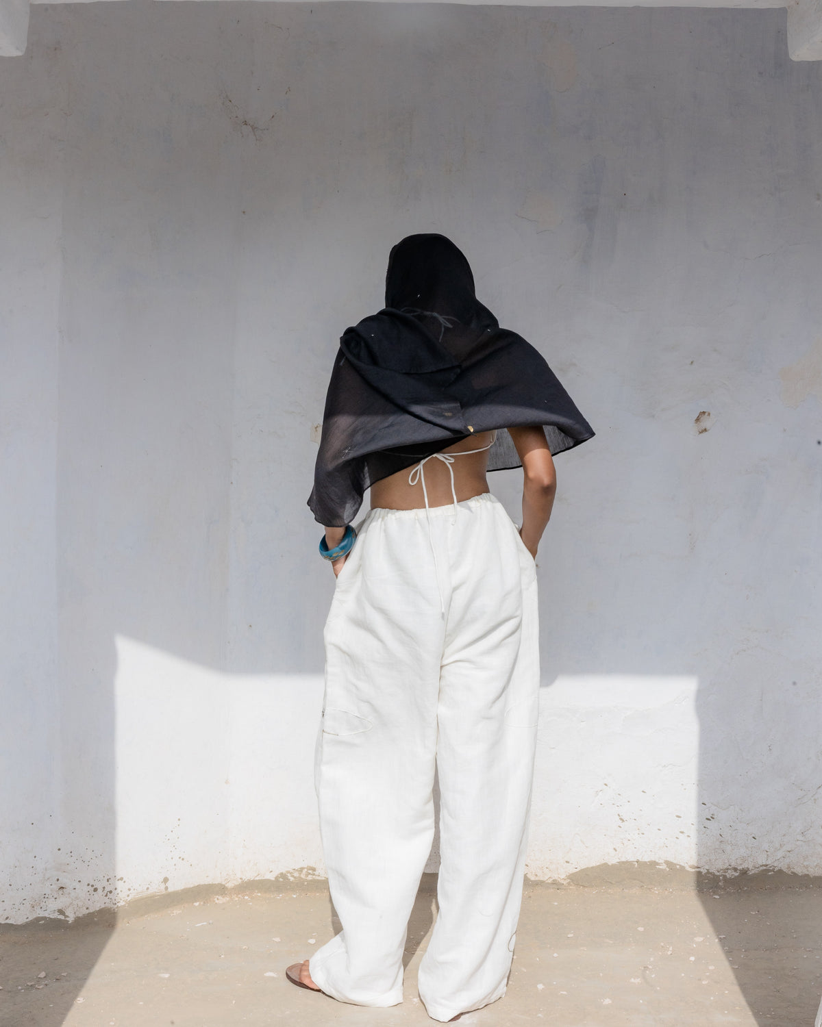 A person standing with their back to the camera in a minimalist white-walled room, wearing a loose white linen cotton trousers with drawstring  and handwork.