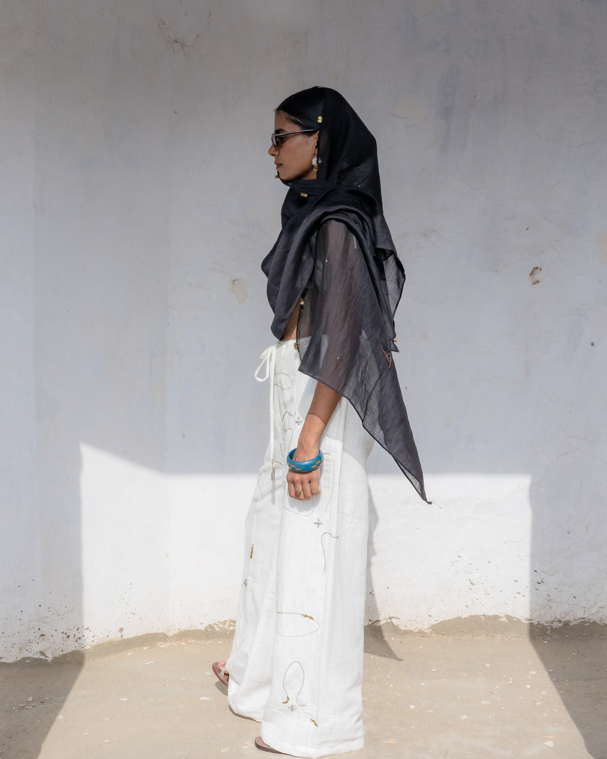 A person wearing a black headscarf and sunglasses, styled with white wide-leg pants, standing in a minimalist white-walled corridor.