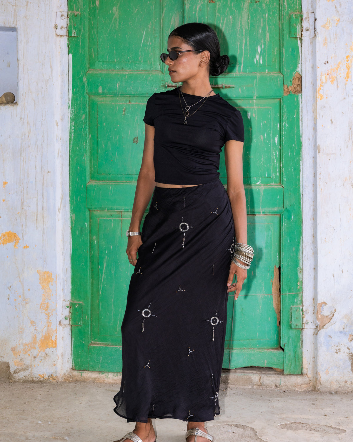 A woman wearing a black two-piece outfit consisting of a short-sleeve crop top and a long skirt with silver keyhole circle embroidered details, standing in front of a weathered green door.