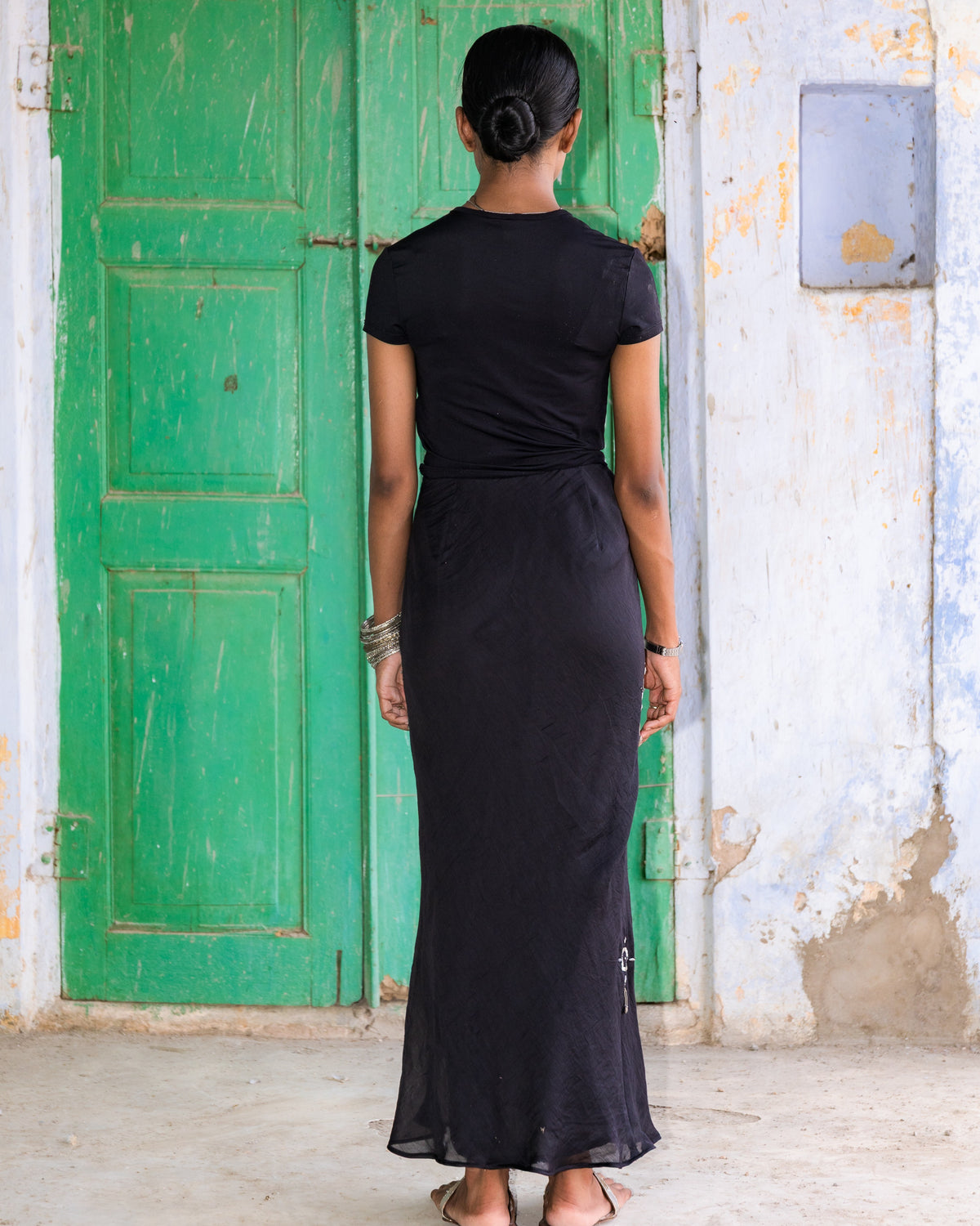 A woman viewed from behind wearing a chanderi black midi skirt, standing in front of a weathered green wooden door.