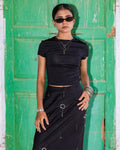 A model wearing a black short-sleeve crop top and matching black skirt with silver circular details, styled with sunglasses, layered necklaces, and bracelets, standing in front of a weathered green wooden door.