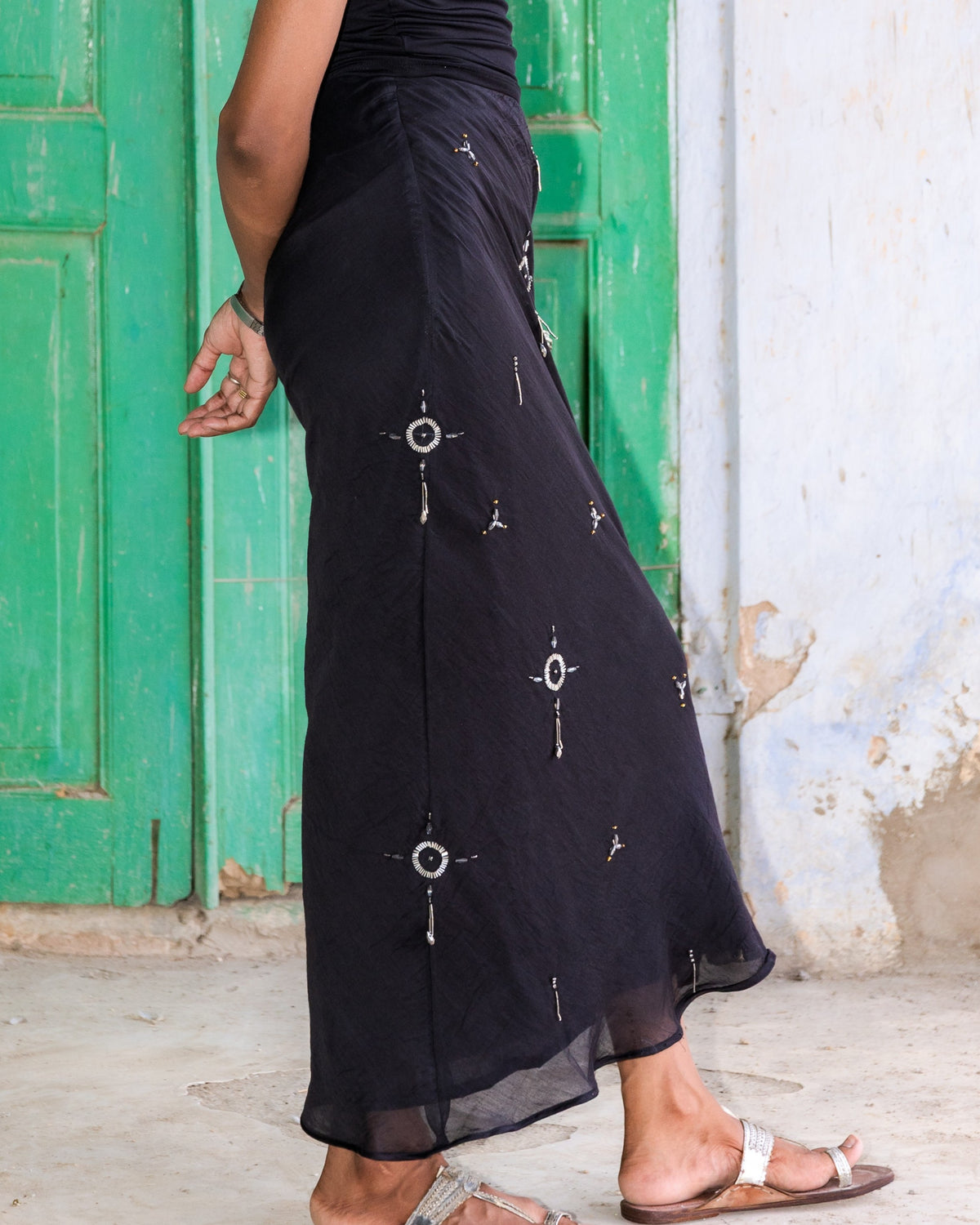 A woman in profile wearing a black midi skirt cut on bias with side zipper with silver beads embroidery, standing in front of a weathered green door and white wall.