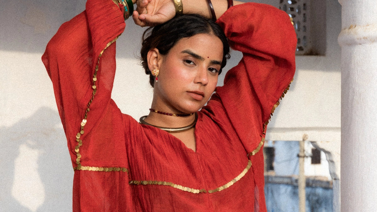 A woman wearing a flowing rust red traditional garment with gold sequence and multiple gold bangles, posing with her arms raised against a white architectural background.