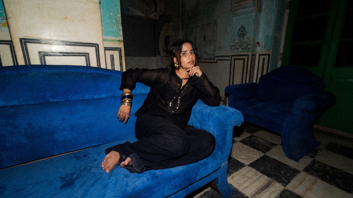 A woman in a black embellished outfit sits barefoot on a large blue velvet sofa in a dimly lit room of an old Indian Haveli.
