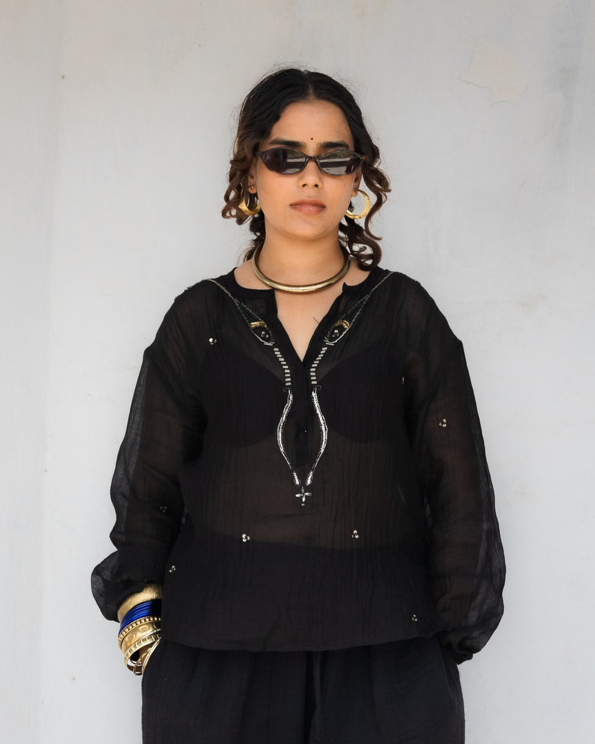 Woman wearing a black long-sleeve sheer kurta with detailed beadwork around the placket and black pants stands inside a white-walled architectural opening.