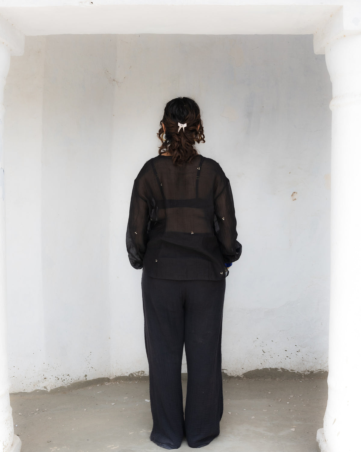A person viewed from behind wearing a black sheer long-sleeve top, minimal beadwork and black stright pants, standing inside a white-walled architectural alcove.