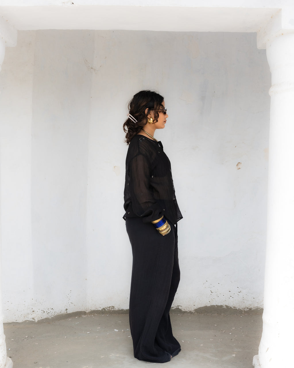 A woman in a black sheer long-sleeve kurta with hand bead work and black wide-leg trousers stands in profile.