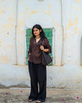 A woman stands wearing a dark brown long-sleeve semi sheer embellished top, black long pants, and carrying a  shoulder bag.