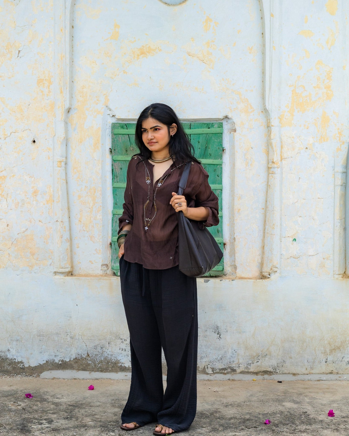 A woman stands wearing a dark brown long-sleeve semi sheer embellished top, black long pants, and carrying a  shoulder bag.